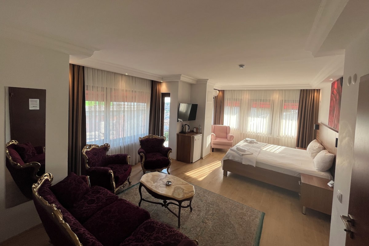 A hotel room with a bed, two armchairs, a sofa, a small table, a TV, and a window with curtains.