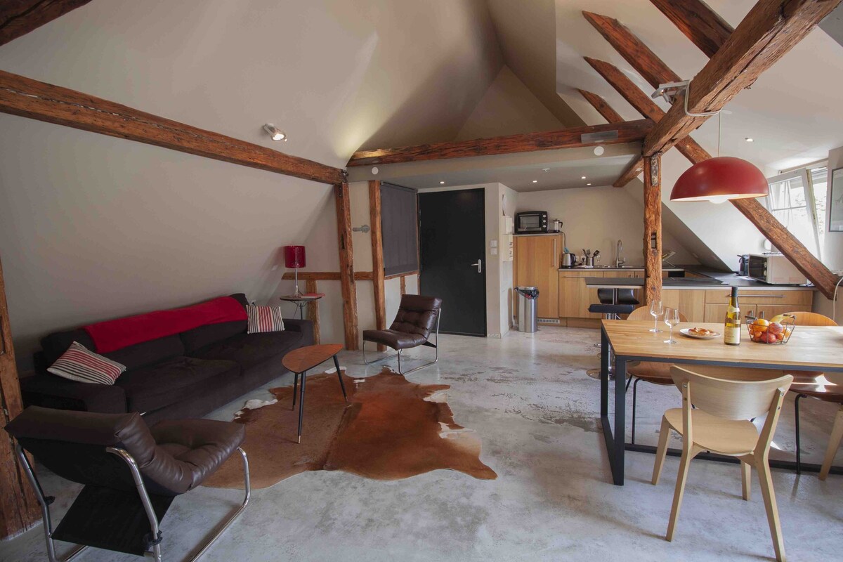 Ein geräumiges Loft mit freiliegenden Holzbalken, einem Essbereich mit roter Pendelleuchte, einem Ledersofa und einem Kuhfellteppich.
