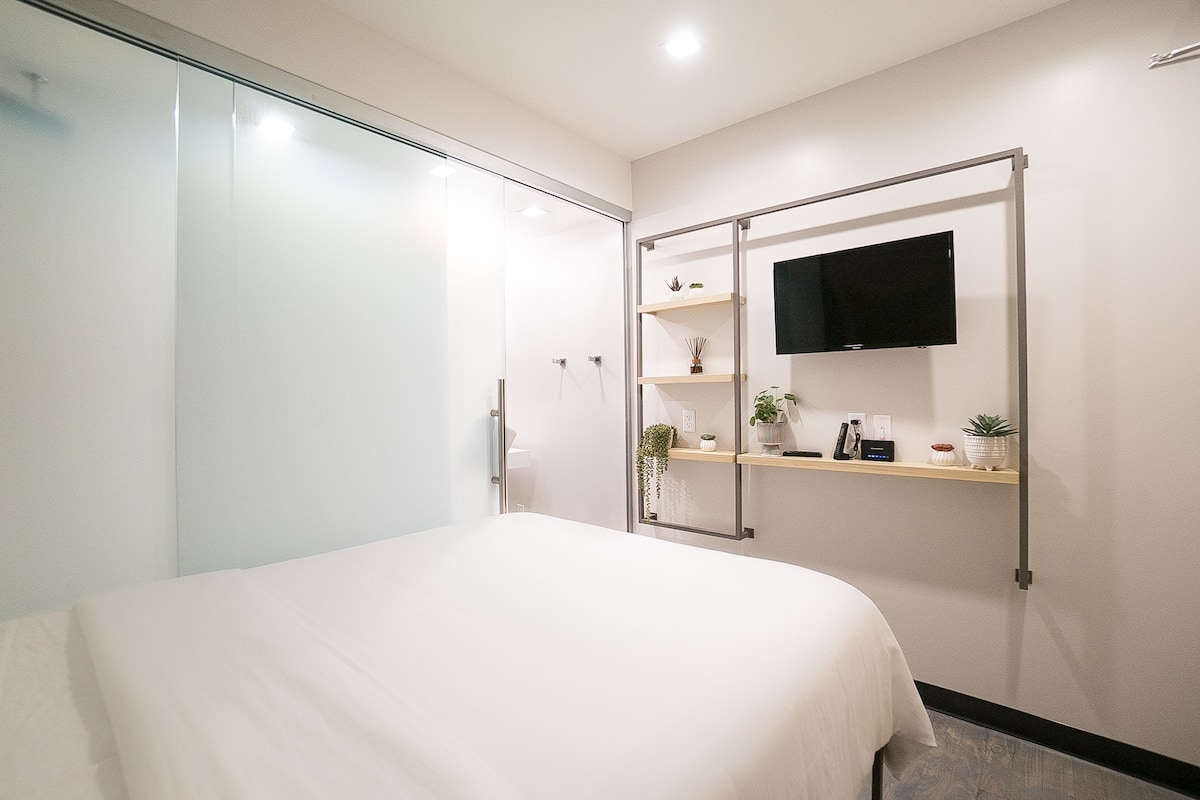 A modern hotel room with a bed, a glass shower enclosure, a mounted TV, and a shelf with decorative items.