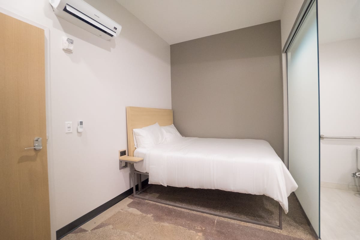 A small, neatly arranged hotel room with a single bed, a bedside table, a wall-mounted air conditioner, and a door.