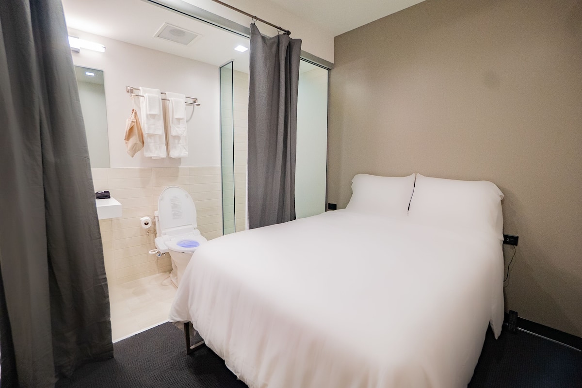 A hotel room with a bed, a bathroom with a toilet and sink, and a glass shower enclosure.