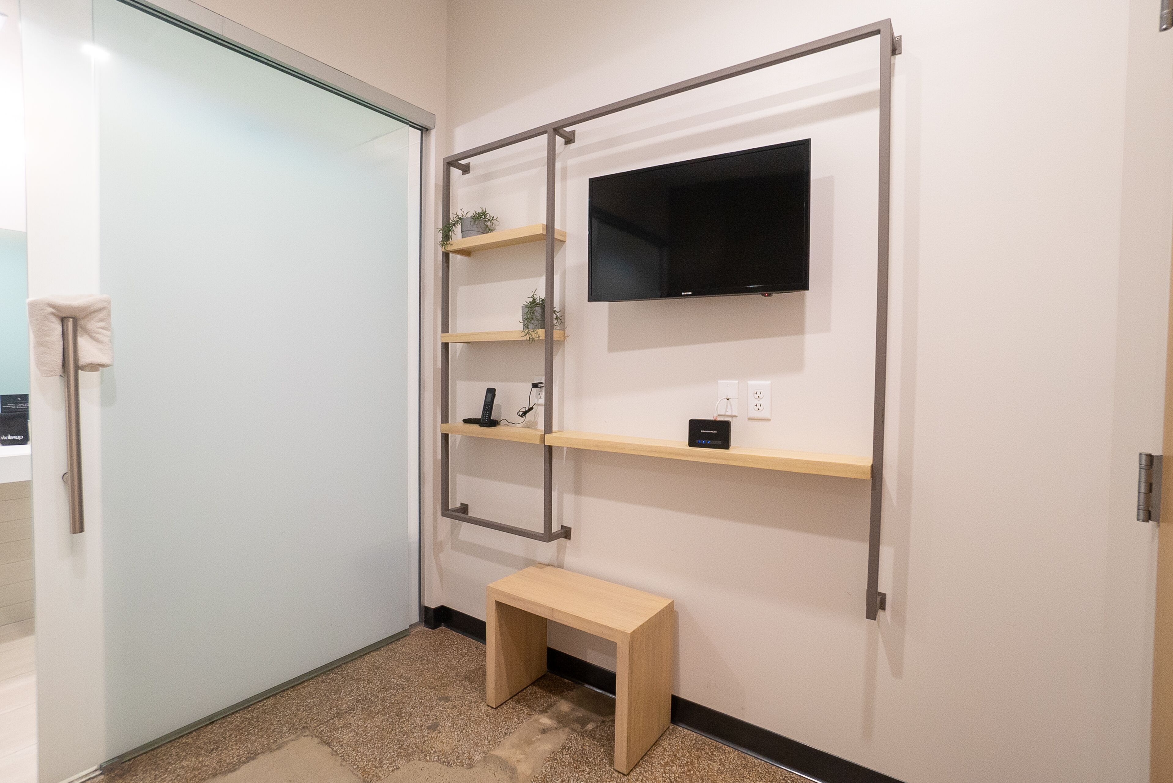 A bathroom with a glass door, a wooden bench, and a wall-mounted TV.