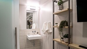 A modern bathroom with a white sink, mirror, and towel rack. There are wooden shelves with plants and a remote control.