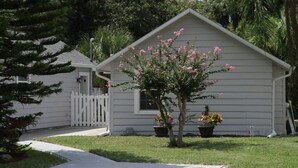 A white single-story house with a front porch, a small tree with pink flowers, and two potted plants. - 8th Green Cottage "A relaxing Oasis in the city" Guesthouse (Orlando)