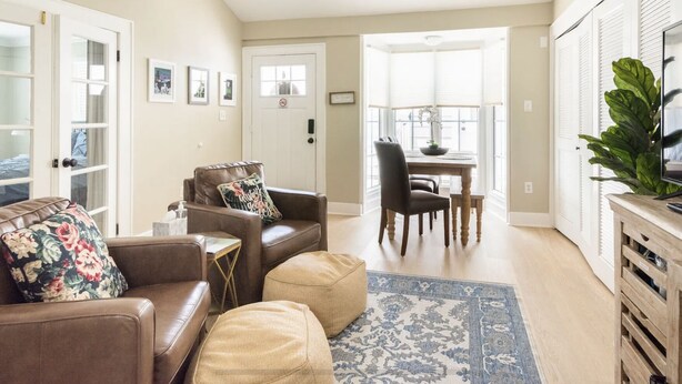 A living room with a brown leather sofa, a beige ottoman, a dining table with chairs, and a TV on a wooden stand. - 8th Green Cottage "A relaxing Oasis in the city" Guesthouse (Orlando)
