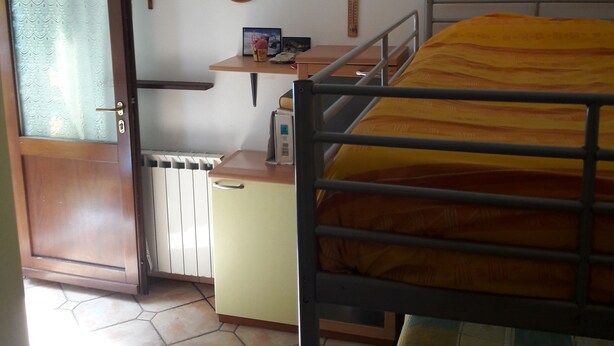 Een kleine, compacte kamer met een stapelbed, een bureau met stoel, een radiator, een houten deur en een wandplank met enkele spullen.