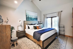 A bedroom with a wooden bed, a nightstand, a small stool, a wall-mounted lamp, and a large window with curtains.