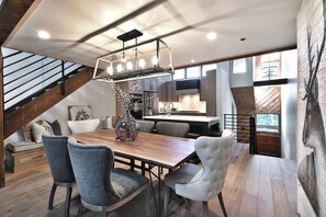 Une cuisine moderne dotée d’appareils électroménagers en acier inoxydable, un coin repas avec une table en bois et des chaises rembourrées, et un escalier avec des poutres apparentes.