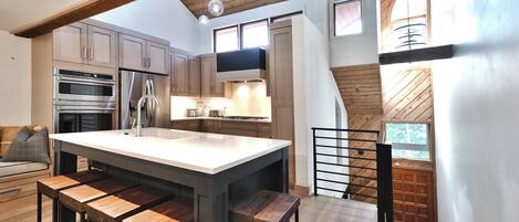 Una cocina moderna con gabinetes de madera, una isla grande con taburetes de bar, electrodomésticos de acero inoxidable y vistas al exterior a través de una ventana.