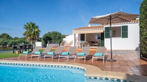 A poolside area with lounge chairs and a covered seating area, a clear blue sky, and a white building with green shutters.