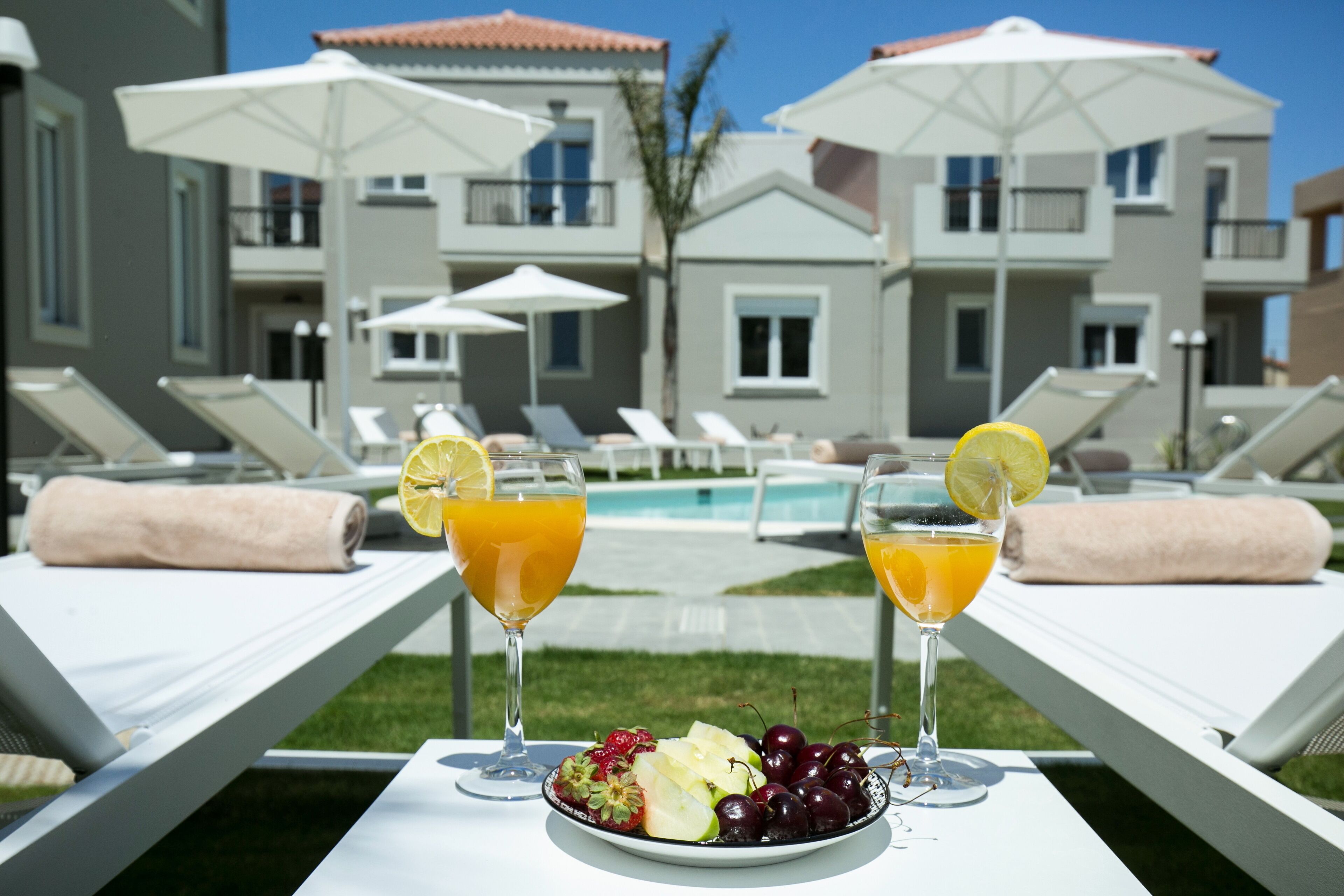 Une table placée au bord de la piscine, avec deux boissons et une assiette de fruits, entourée de chaises longues et de parasols.