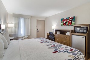 A hotel room with a large bed, a desk, a chair, a television, and a refrigerator. - Red Roof Inn St George, SC (St. George)