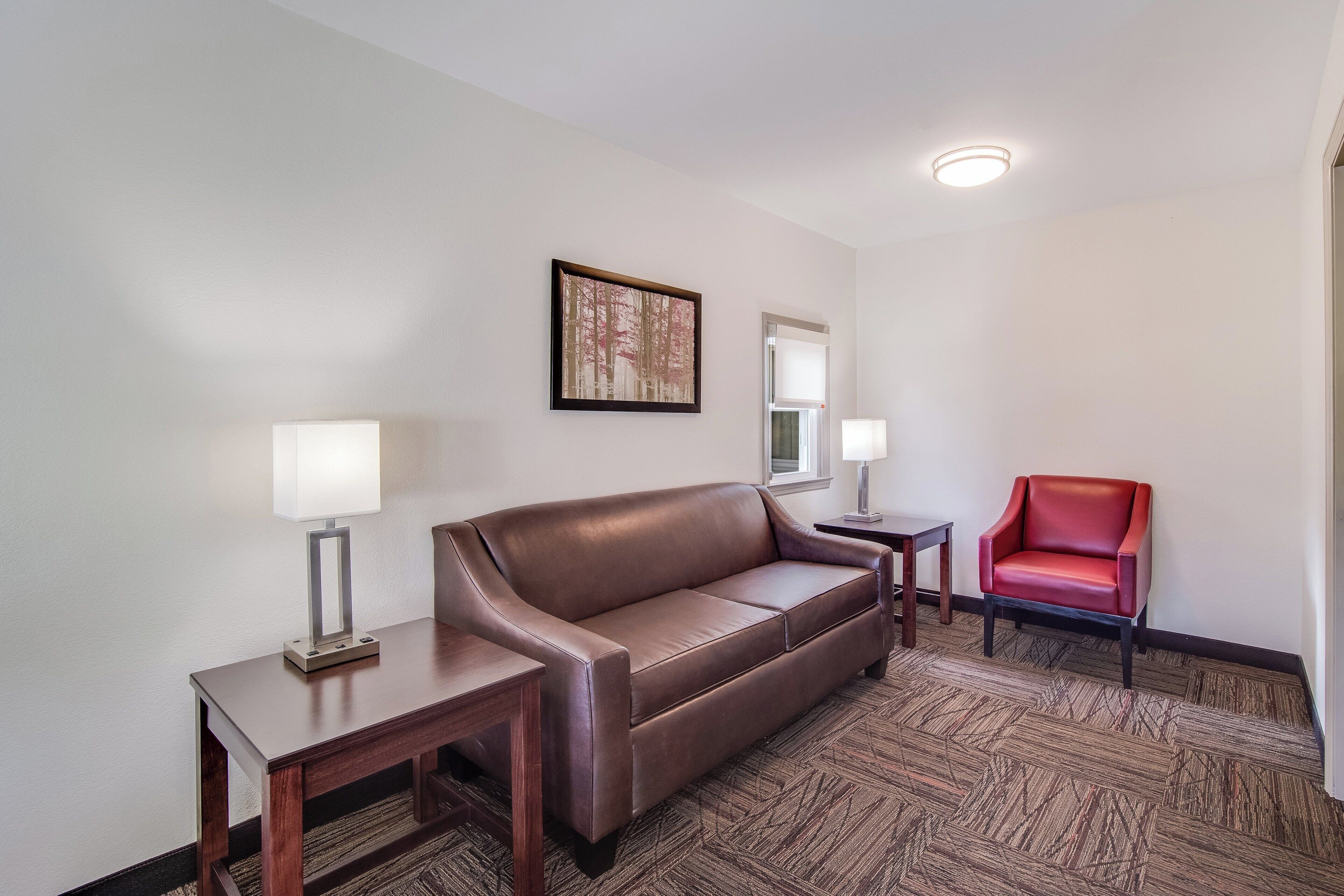 A hotel room with a brown leather sofa, two bedside tables with lamps, a red chair, and a framed picture on the wall.