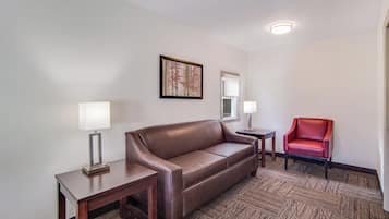 A hotel room with a brown leather sofa, two bedside tables with lamps, a red chair, and a framed picture on the wall.