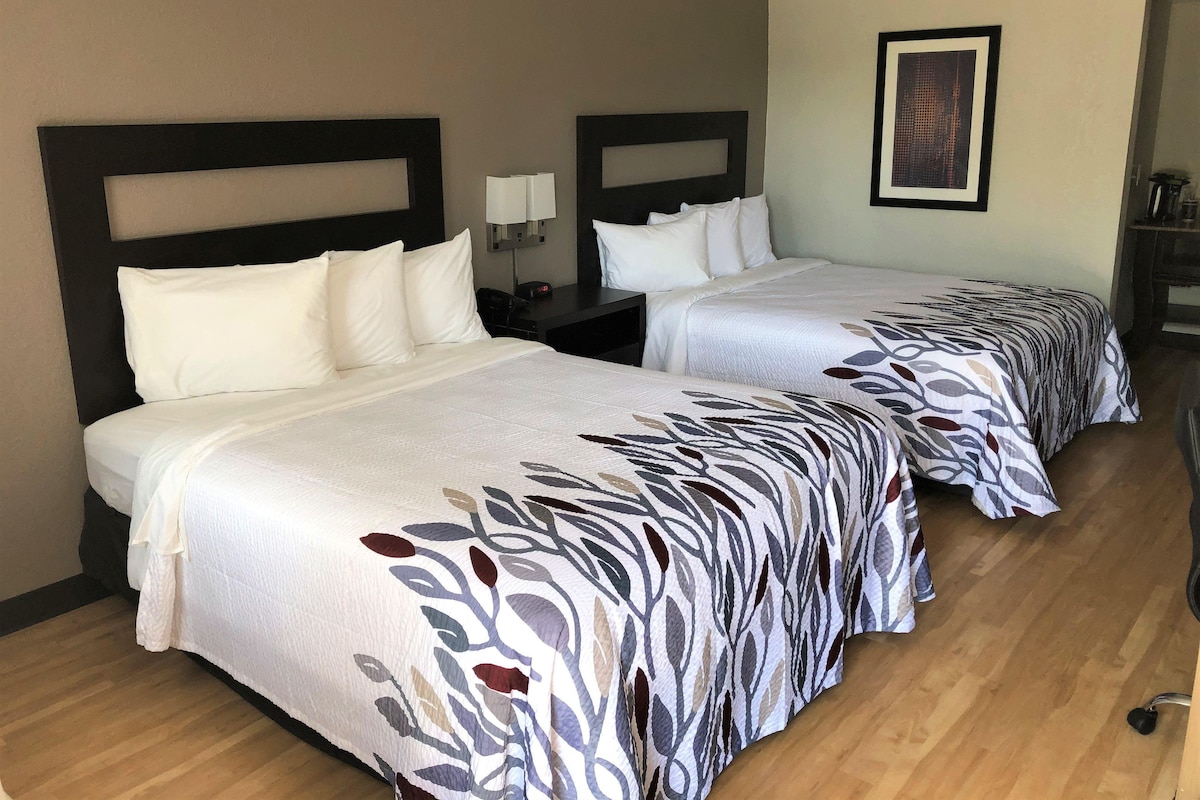 Une chambre d’hôtel avec deux lits, chacun doté d’une tête de lit, de draps blancs et d’une couvre-lit à motifs. On y trouve une table de chevet avec une lampe, ainsi qu’un tableau encadré accroché au mur.