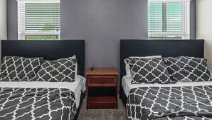 A bedroom with two beds, each with a black and white patterned bedspread, a small wooden nightstand with a drawer, and a window with white blinds.
