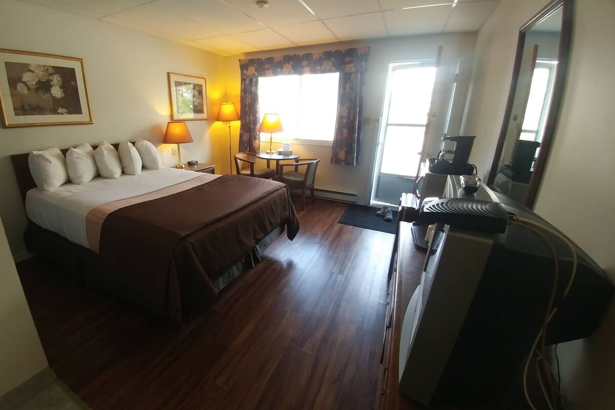 A hotel room with a bed, two bedside lamps, a small table with a chair, a large mirror, and a window with curtains.