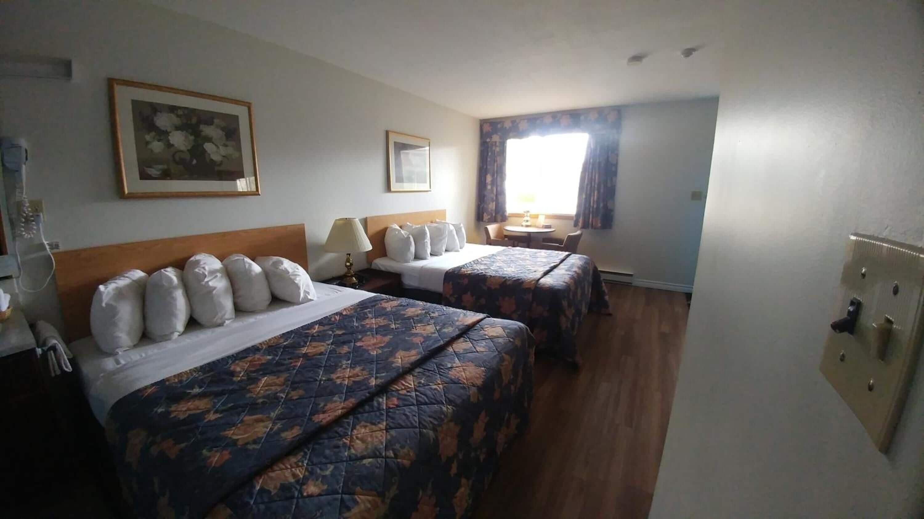 Habitación de hotel con dos camas, una mesita, una silla, una lámpara y un ventanal con cortinas.