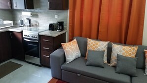 Een compacte woonruimte met kitchenette, grijze bank met gekleurde kussens en oranje gordijnen.