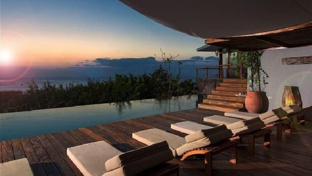 Ein unendlicher Pool mit Holzterrasse und Liegestühlen, mit Blick auf eine Berglandschaft bei Sonnenuntergang.