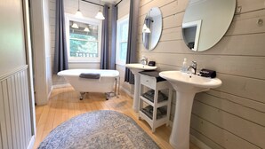 A bathroom with a freestanding bathtub, two sinks, a mirror, and a window with curtains. - Deluxe Room Two Queen Beds (Dahlonega)