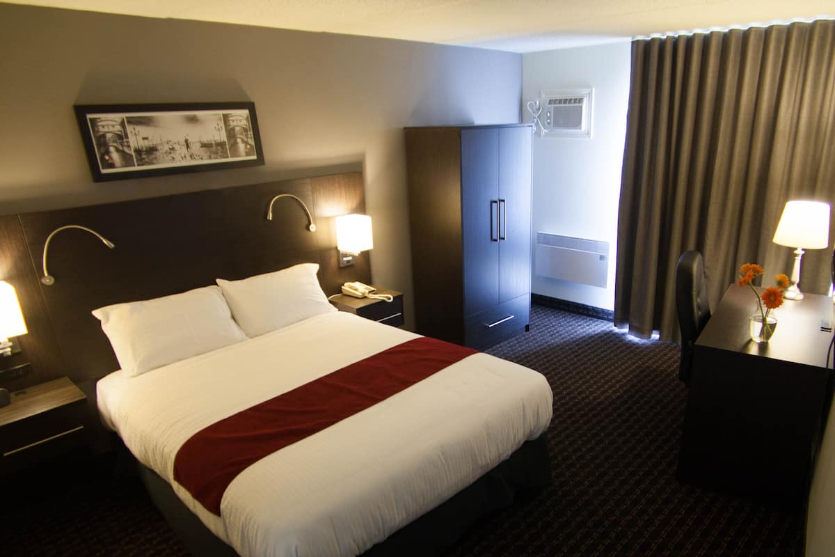 A hotel room with a large bed, two bedside tables with lamps, a wardrobe, a desk with a lamp, and a window with curtains.