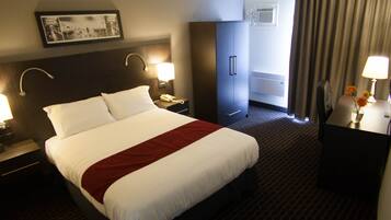 A hotel room with a large bed, two bedside tables with lamps, a wardrobe, a desk with a lamp, and a window with curtains.