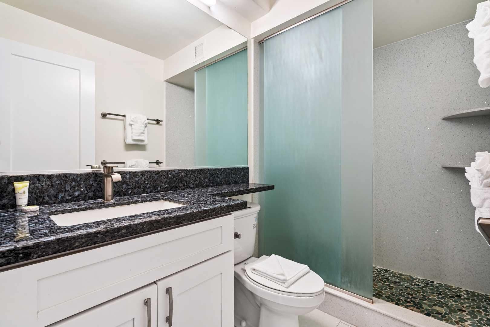 Een badkamer met een granieten aanrechtblad, witte kasten, een wit toilet en een douche met een groene glazen deur.
