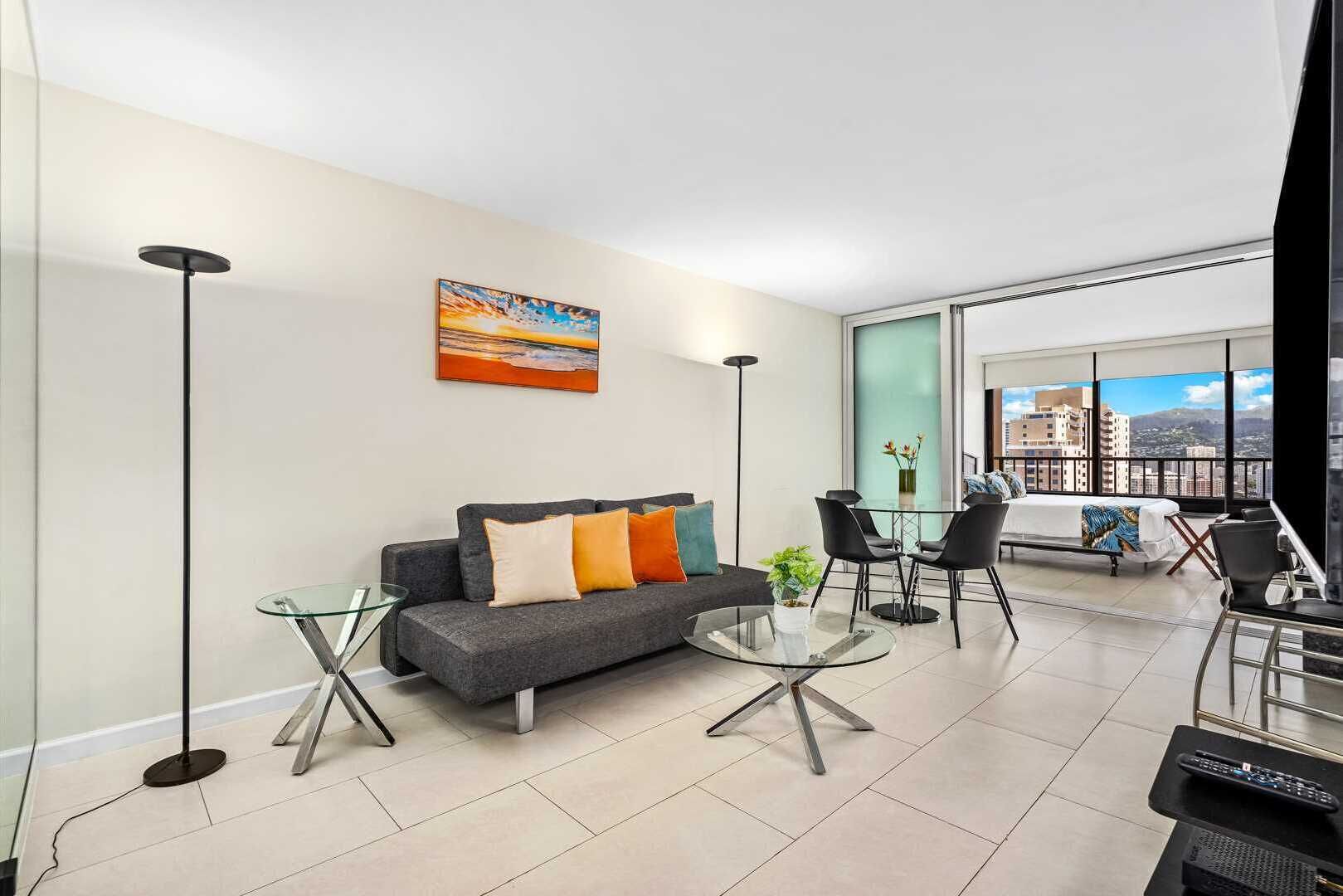 A modern living room with a gray sofa, a glass coffee table, and a dining area with black chairs. A painting hangs on the wall, and the room has large windows with a city view.