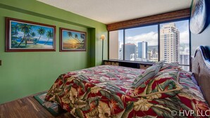 A hotel room with a large bed, two tropical-themed paintings on the wall, a city view through the window, and a lamp beside the bed.
