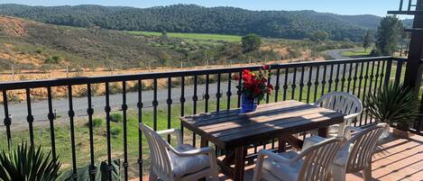 A balcony with a table and chairs, a vase with flowers, and a view of a river and hills.