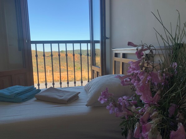 A room with a bed, a window with a view of a desert landscape, and a vase with flowers.