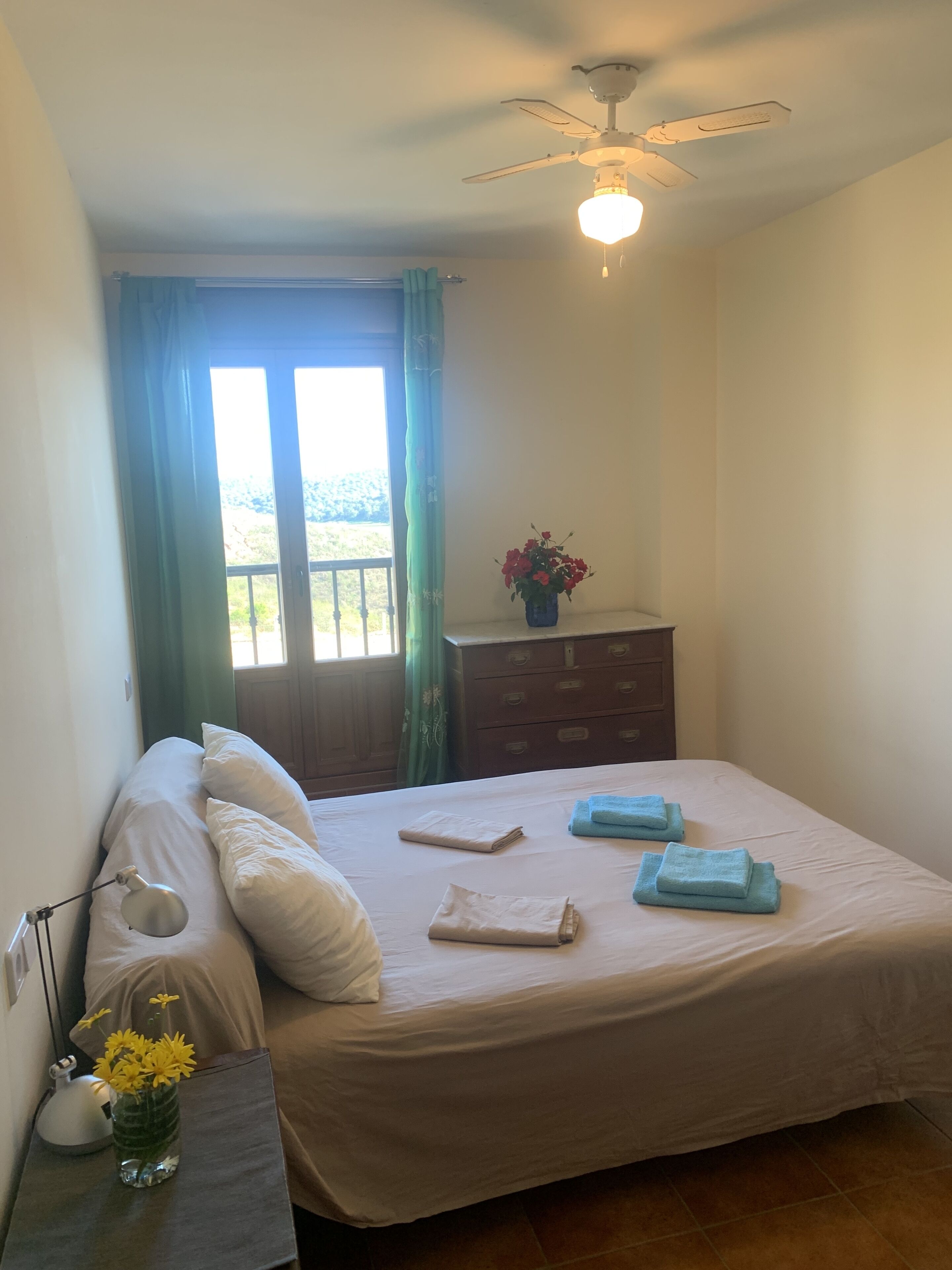 Un dormitorio con cama, mesita de noche, cómoda, ventilador de techo y ventana con cortinas.
