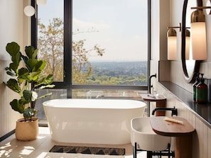 A modern bathroom with a freestanding bathtub, a small wooden table with toiletries, and a large window offering a city view.