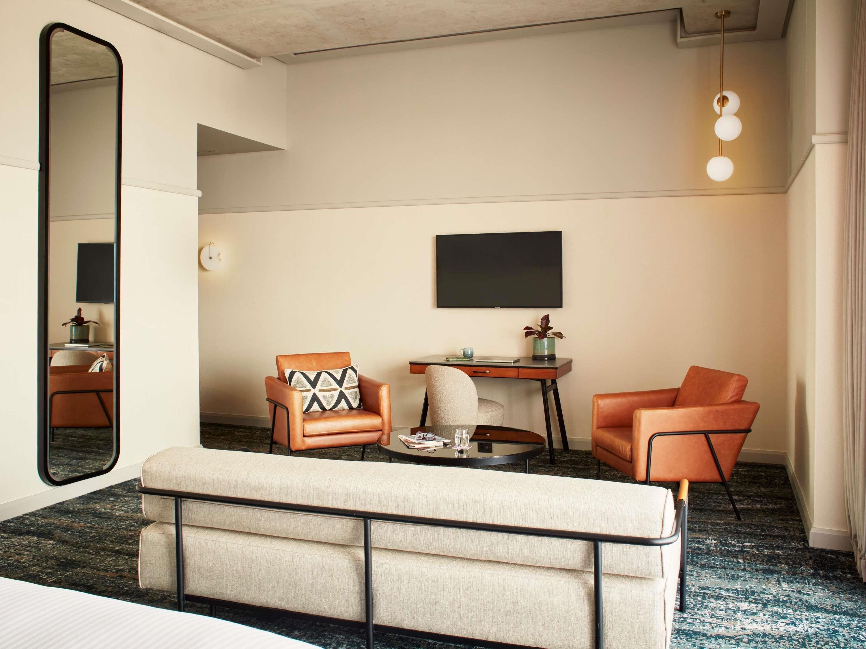 A hotel room with a large mirror, a sofa, two armchairs, a small table with a glass, and a TV on the wall.