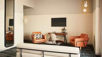 A hotel room with a large mirror, a sofa, two armchairs, a small table with a glass, and a TV on the wall.