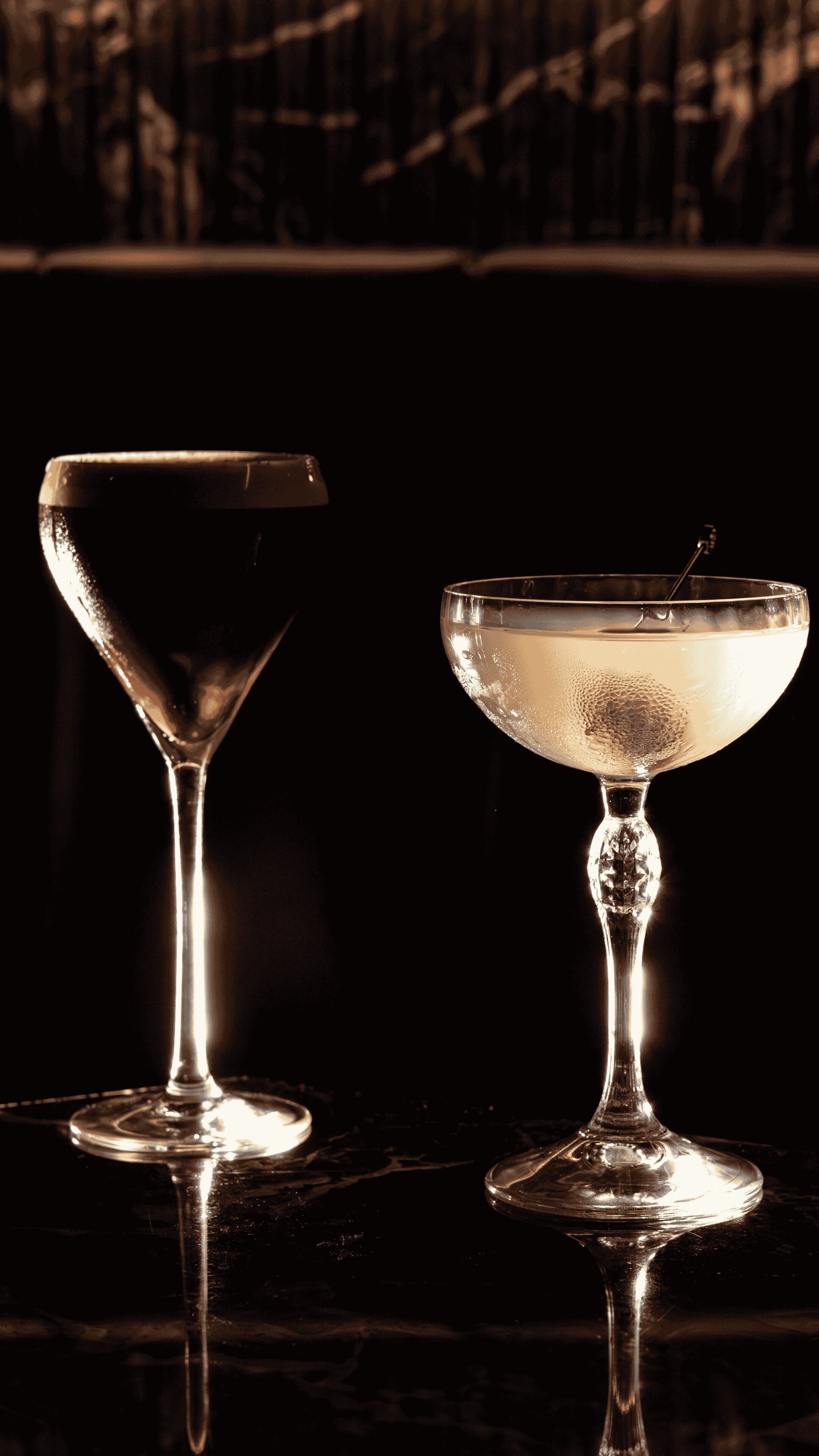 Two stemmed glasses on a reflective surface with a dark background.
