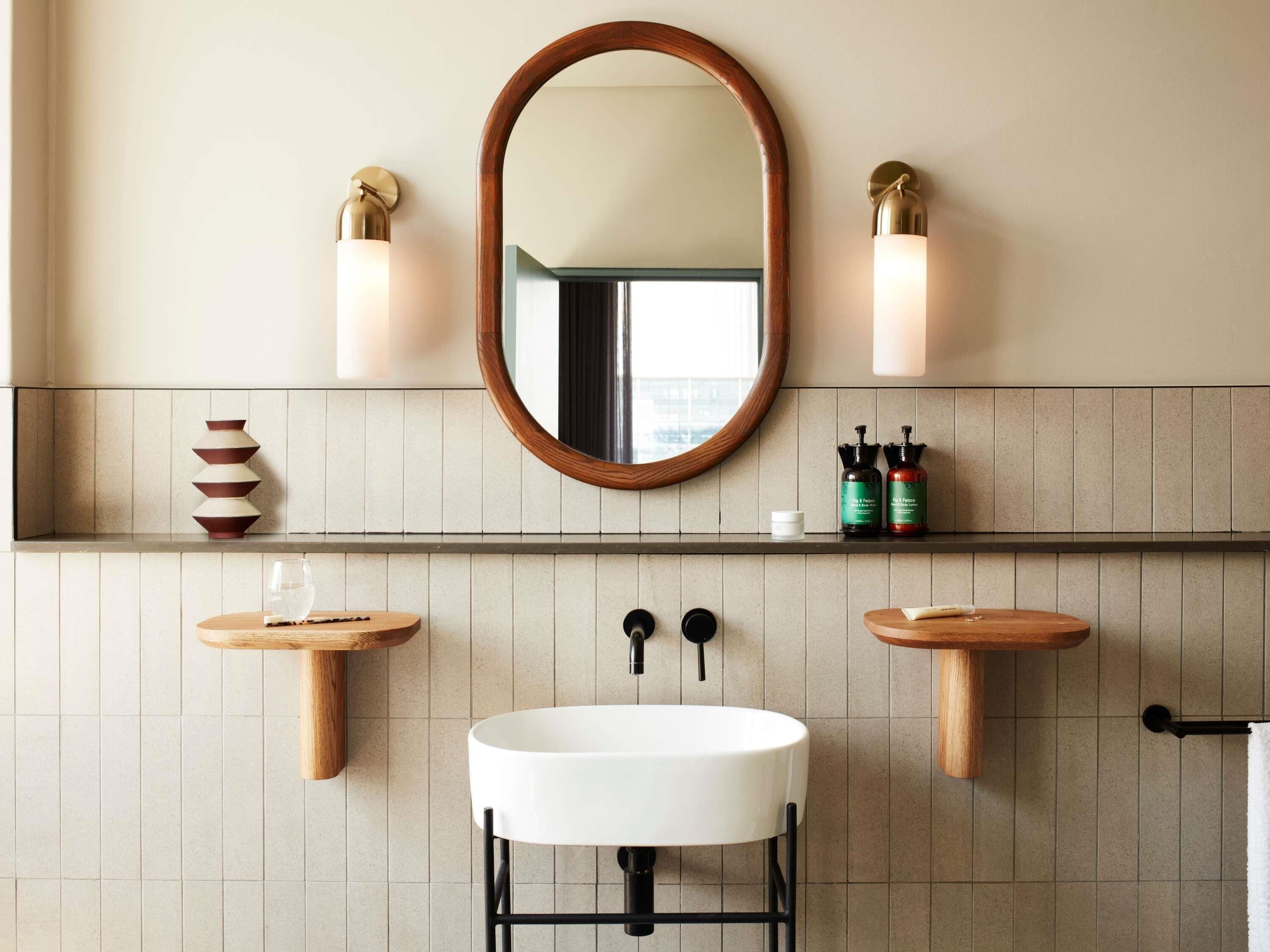 Un baño con un espejo redondo, dos luces empotradas, un lavabo blanco y estantes de madera con artículos de aseo.