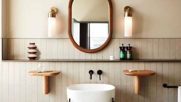 Un baño con un espejo redondo, dos luces empotradas, un lavabo blanco y estantes de madera con artículos de aseo.