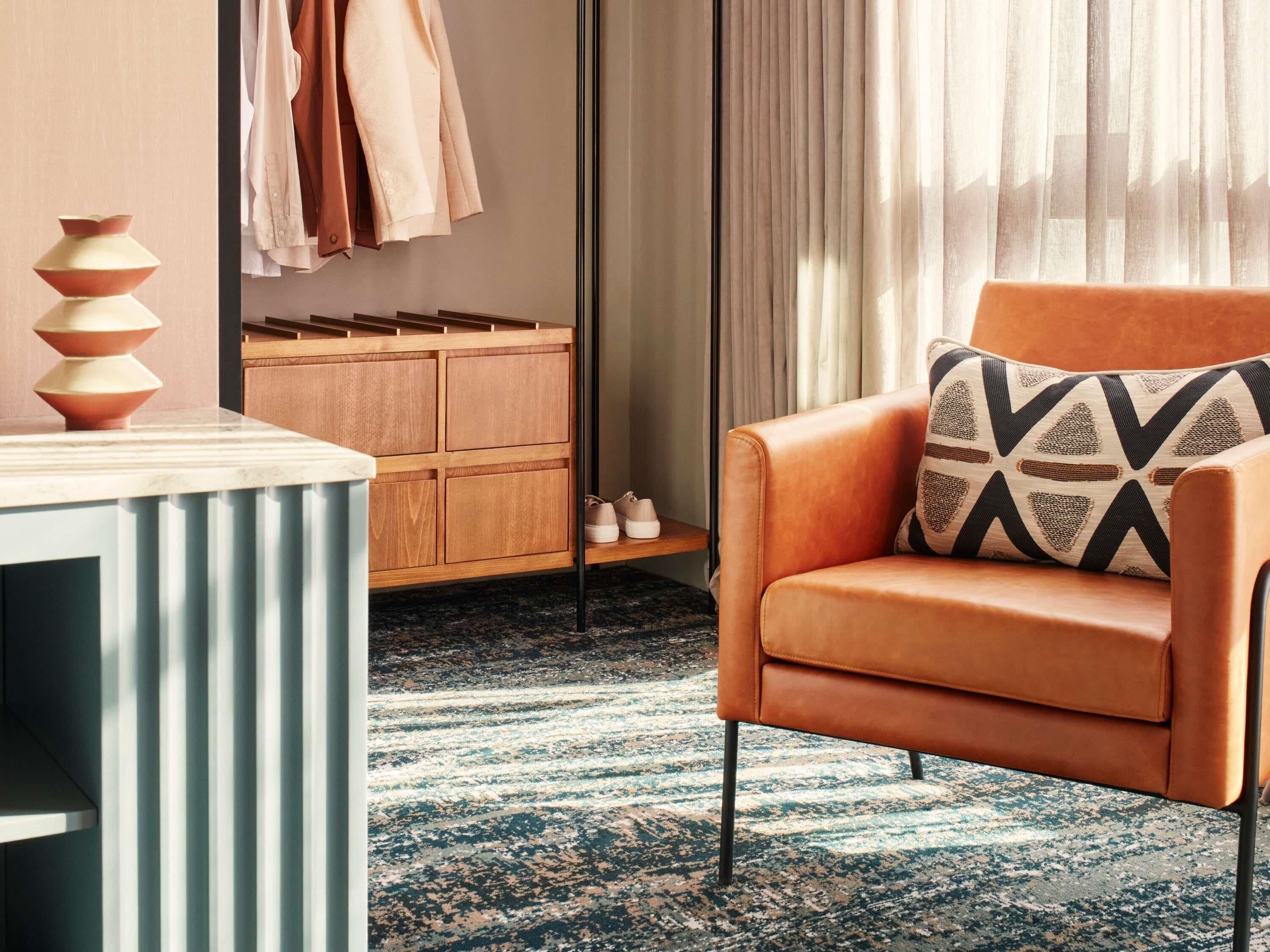 Una habitación de hotel moderna con un aparador de madera, un sillón de cuero con cojines estampados y una mesa de mármol.