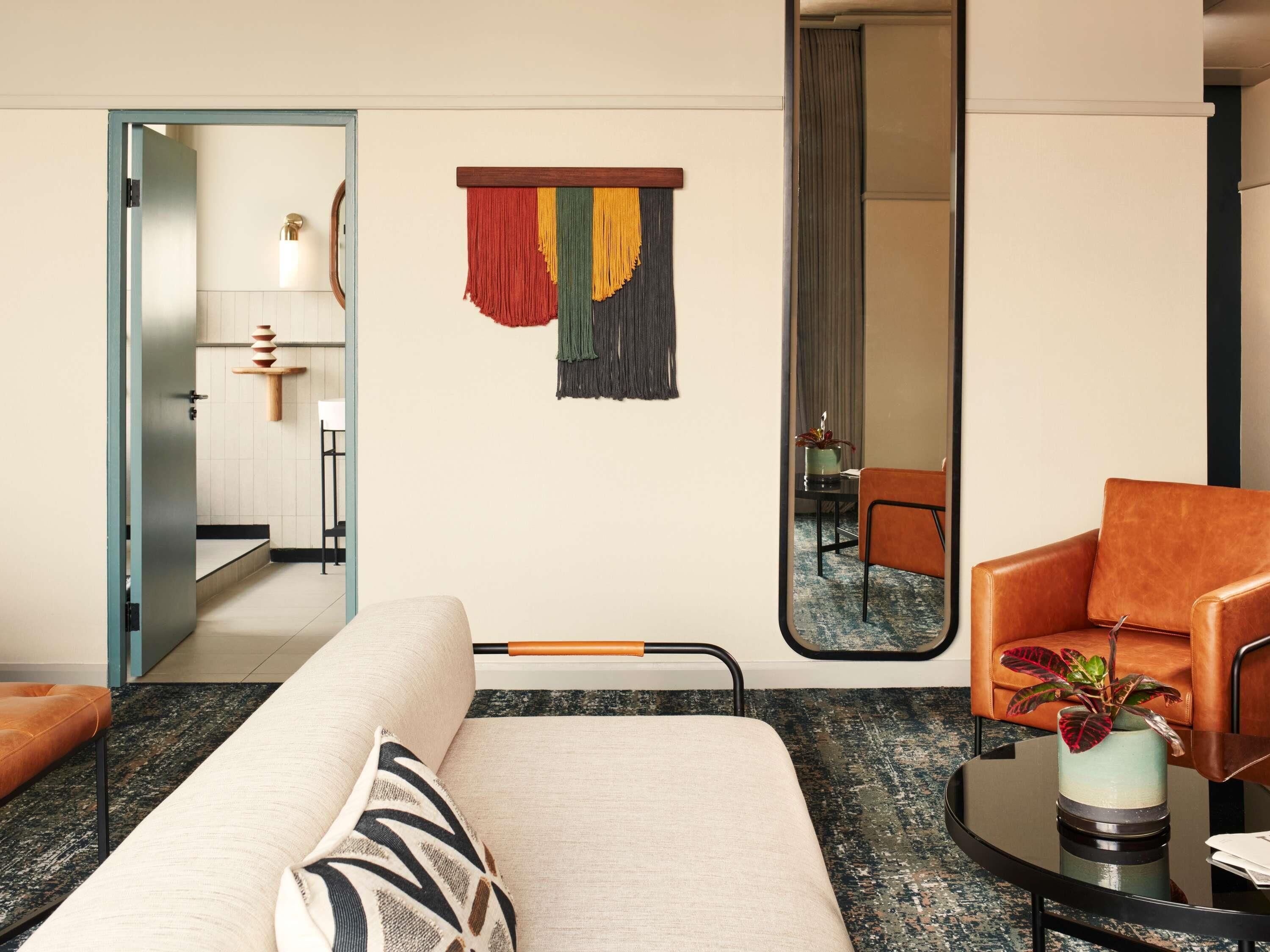 A modern hotel room with a sofa, a chair, a mirror, and a decorative wall hanging.