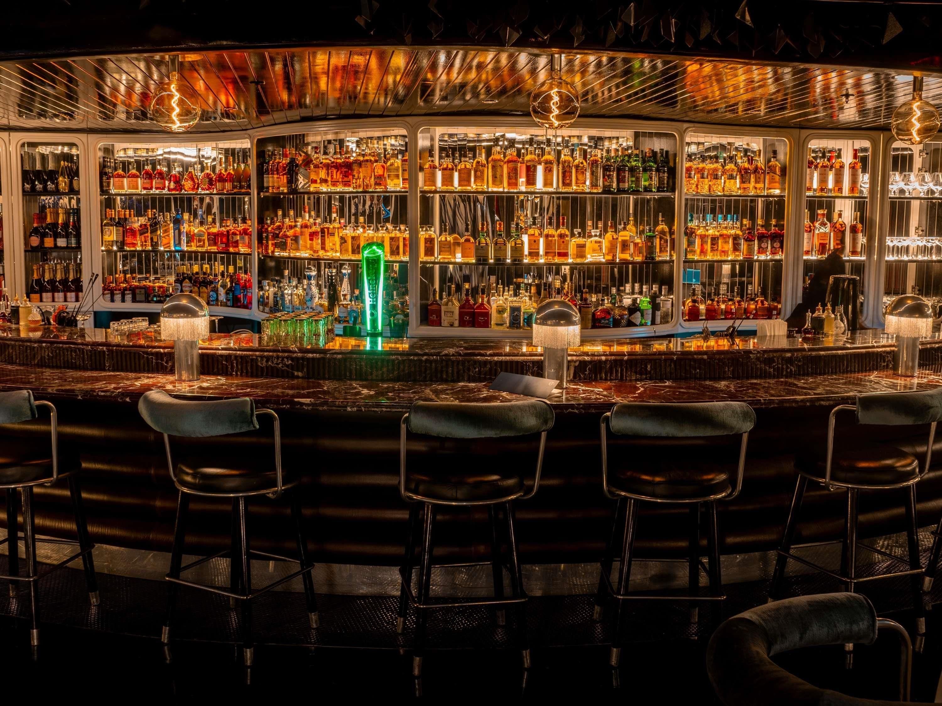 Un bar con múltiples estantes llenos de botellas de licores, taburetes y una encimera de mármol.