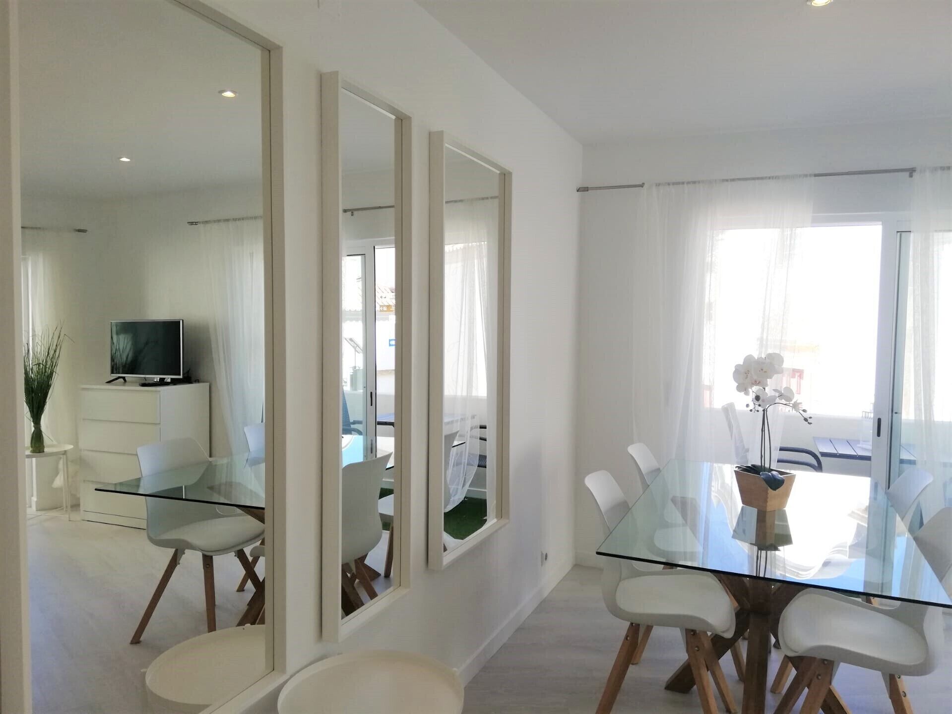A modern dining area with a glass table and chairs, a television in the background, and large windows with sheer curtains.