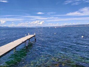 Un muelle de madera se extiende sobre aguas tranquilas y cristalinas, donde se distinguen veleros a lo lejos y una costa distante.