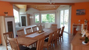 A dining area with a wooden table and chairs, a kitchen in the background, and large windows allowing natural light.