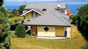 A yellow house with a grey roof, surrounded by greenery and a view of a body of water and distant hills.