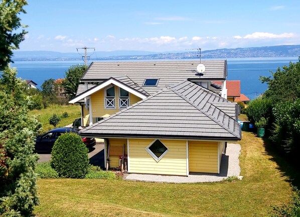 Una casa amarilla con techo gris, rodeada de vegetación y vistas a un cuerpo de agua y colinas distantes.