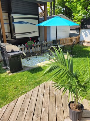 A patio area with a grill, a table with chairs, a blue umbrella, and a wooden deck. - The Big Happy Place (Fergus)
