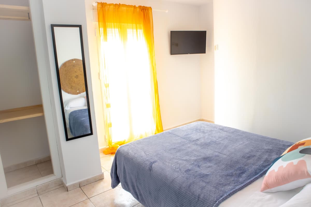 Un dormitorio con cama, espejo, televisión y ventana con cortinas.