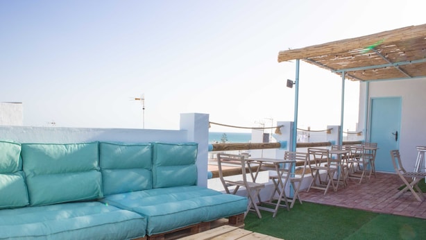 A rooftop terrace with a wooden table and chairs, a blue sofa, and a view of the sea. - Hostal Salitre (Conil de la Frontera)